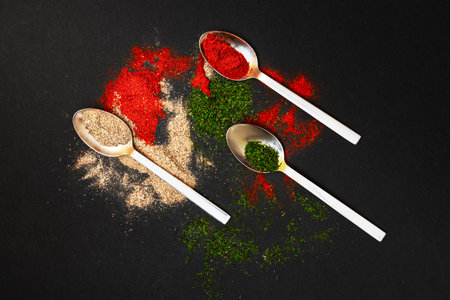 Loose spices in metal spoons are beautifully scattered against a black background, creating a...