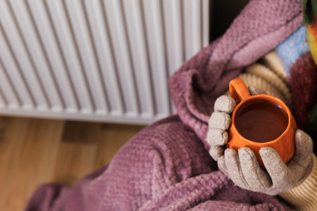 A close-up of a girl holding a cup of tea. She sits near a radiator, She's wearing a scarf, gloves, and a sweater. She's wrapped in a purple blanket. She's trying to keep warm in a poorly heated roomの写真素材
