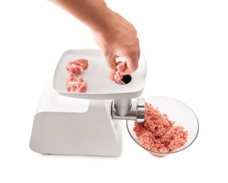 A hand putting raw meat pieces into a white electric meat grinder, producing fresh minced meat into a glass bowl, isolated on white. High quality photoの写真素材