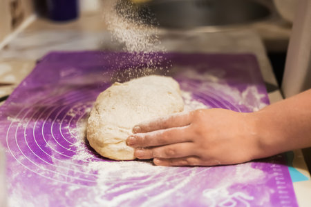 A hand dusting a portion of raw dough with a cloud of flour on a bright purple, floured silicone mat, showing the start of baking preparation. High quality photoの写真素材