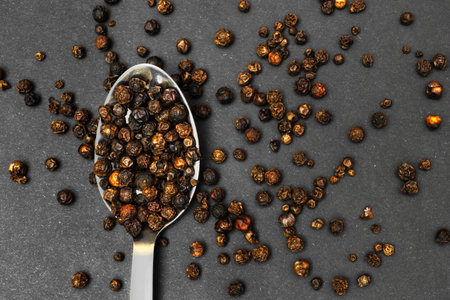 An overhead close-up of a silver spoon overflowing with whole black peppercorns scattered across a dark gray textured surface. High quality photoの写真素材