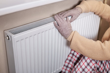 A girl in gloves warms her hands on a barely warm radiator. She wears a cozy sweater and sits on a checkered blanket, indicating a chilly indoor environment.の写真素材