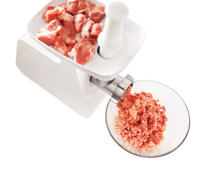 A white electric meat grinder mincing raw pork chunks into ground meat, with the output collecting in a clear glass bowl. High quality photoの写真素材