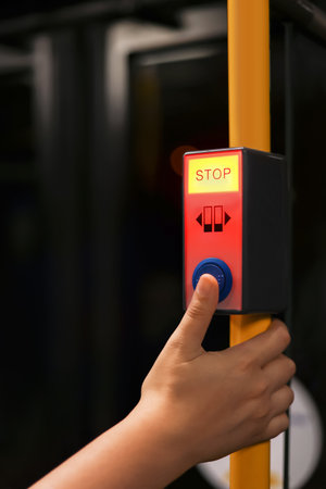A young girl presses the stop button on a city bus. The button is illuminated in red with the word...