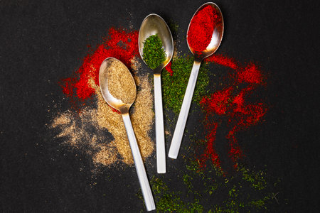 Loose spices in metal spoons are beautifully scattered against a black background, creating a contrast between the colors of red paprika, green dill, and yellow garlic.Top viewの写真素材