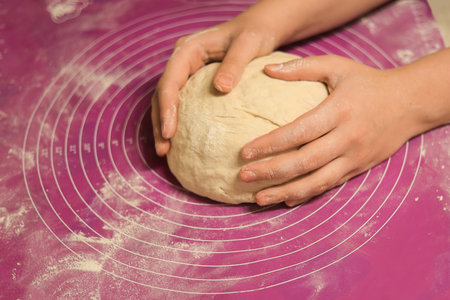 A close-up of hands gently shaping and rounding a large ball of fresh dough on a pink, flour-dusted silicone baking mat. High quality photoの写真素材