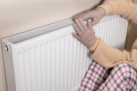 A girl in gloves warms her hands on a barely warm radiator. She wears a cozy sweater and sits on a checkered blanket, indicating a chilly indoor environment.の写真素材