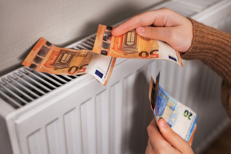 A girl in a sweater counts money near a radiator. The room appears cold, emphasizing the need for heating.の写真素材