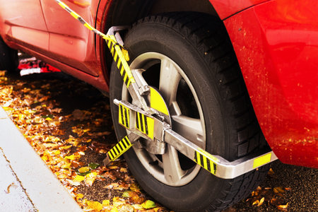 A close-up of a red car's rear wheel fitted with a yellow and black wheel clamp (boot) for a parking violation, on an autumn curb. High quality photoの写真素材