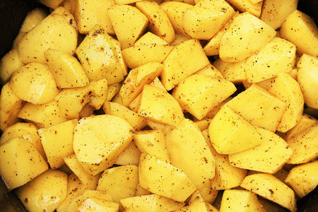 A close-up, full-frame view of raw, diced yellow potatoes seasoned with black pepper and oil inside a black pot. High quality photoの写真素材