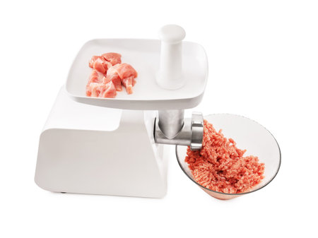 A white electric meat grinder with a white plunger, mincing raw pork chunks into a heap of fresh ground meat in a clear glass bowl on white. High quality photoの写真素材