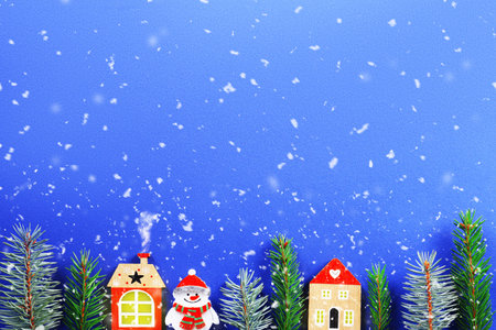 A winter holiday scene featuring wooden toy houses, a snowman, and fir branches under falling snow on a blue background. High quality photoの写真素材