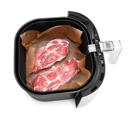 An overhead view of two raw pork neck steaks on parchment paper inside the basket of a black and white air fryer, isolated on white. High quality photoの写真素材