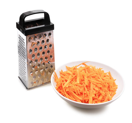 A metal box grater stands next to a white bowl filled with freshly shredded orange carrots, isolated on a clean white background. High quality photoの写真素材