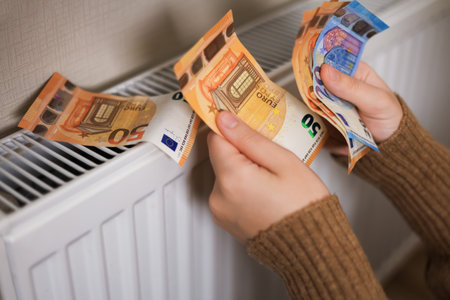 A girl in a sweater counts money by the radiator: euros in 50 and 20 denominations. The room is cold, which emphasizes the need for heating.の写真素材