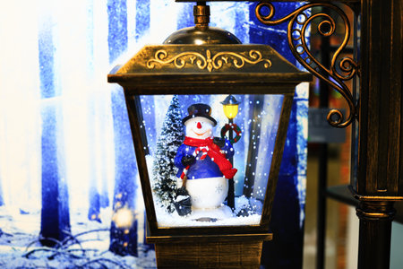 Close-up of a festive snowman figurine inside a decorative glass lantern with fake snow, against a blue winter backdrop. High quality photoの写真素材
