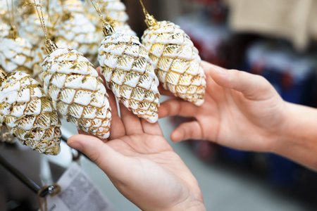 Hands holding and examining white and gold decorative pine cone Christmas tree ornaments hanging in a retail store. High quality photoの写真素材
