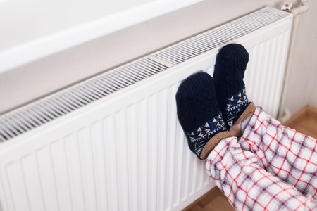 Person wearing cozy patterned slippers and plaid pajamas resting feet on a white radiator to warm up in the cold. High quality photoの写真素材