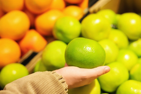 A person's hand holds a large, bright green citrus fruit pomelo over a blurred background of oranges. High quality photoの写真素材