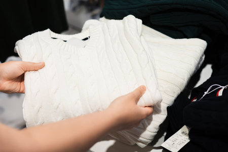 A person's hands hold a folded white cable knit sweater in a clothing store, with other garments visible nearby. High quality photoの写真素材