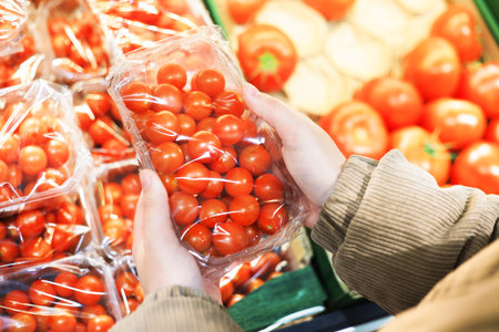 Hands hold a plastic container of bright red cherry tomatoes in a supermarket, surrounded by other fresh produce. High quality photoの写真素材