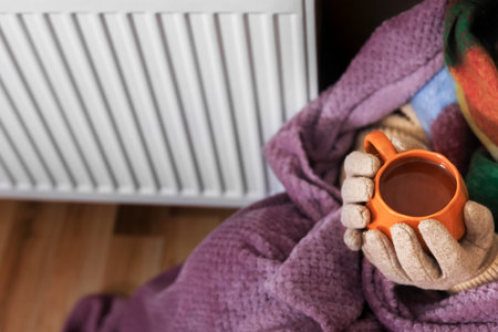 A close-up of a girl holding a cup of tea. She sits near a radiator. She's wearing a scarf, gloves, and a sweater. She's wrapped in a purple blanket. She's trying to keep warm in a poorly heated room.の写真素材