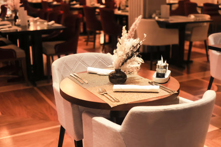 A sophisticated restaurant interior with an intimate table setting, plush chairs, warm lighting, and decorative dried grass. High quality photoの写真素材