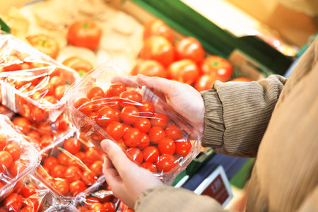 Hands holding a plastic container of bright red cherry tomatoes in the produce section of a grocery store or market. High quality photoの写真素材
