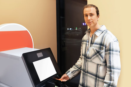 A young man in a plaid shirt uses a self-service terminal, inserting a card, possibly at an airport...