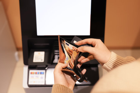 Hands holding a wallet with Euro banknotes and a credit card in front of an ATM screen, ready for a...
