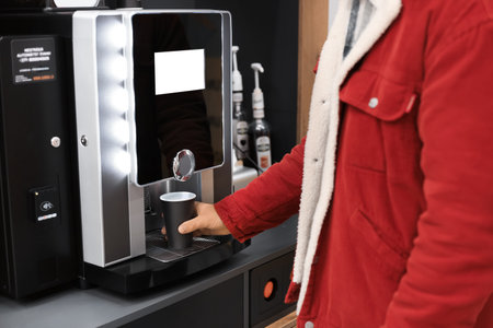 Young man in a red corduroy jacket using a modern, automatic coffee machine to dispense a hot...