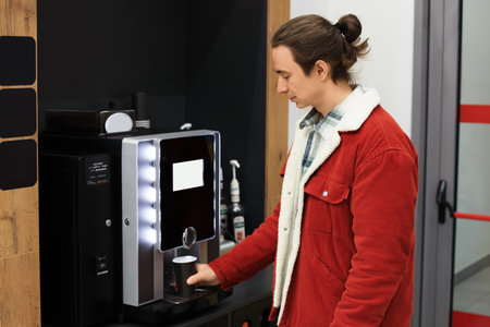 Young man in a red jacket is using an automatic coffee machine in an office or waiting area for a...