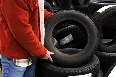 Customer holding a car tire while choosing new wheels in an auto store with stacks of tires. High...