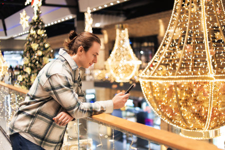 Young man leaning on railing using phone amidst festive Christmas lights and decorations in a shopping mall. High quality photoの写真素材