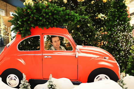 Happy man sitting in a red retro car with a Christmas tree on the roof against a festive background...