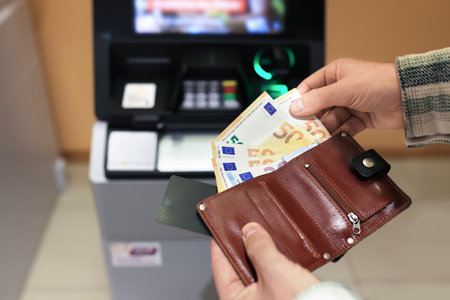 Man hands holding brown wallet with fifty euro banknotes and credit card near ATM machine. High...