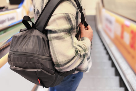 Close up of man with gray backpack standing on escalator. Person wearing plaid shirt in shopping...
