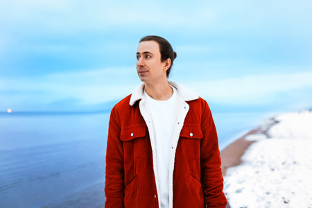 man in a red jacket standing on a snowy beach looking away against a blue sea background. High quality photoの写真素材