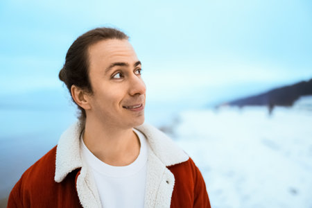 Smiling young man with long hair in red jacket looking away against a snowy winter background.の写真素材