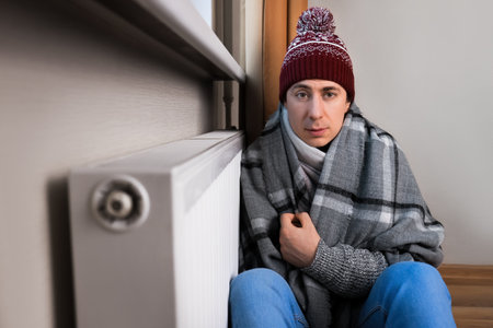 Freezing man in hat and blanket sitting next to cold radiator, struggling to keep warm at home...