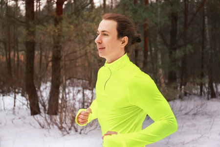 Side view of a male runner with a man bun jogging in a snowy forest. Athlete in a neon yellow shirt...