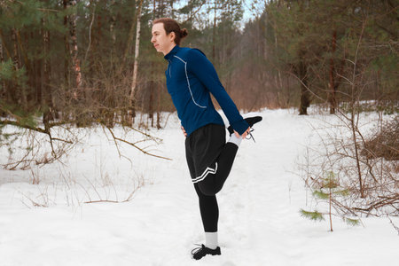 Young male runner stretching leg muscles in a snowy forest. Man warming up before jogging outdoors....