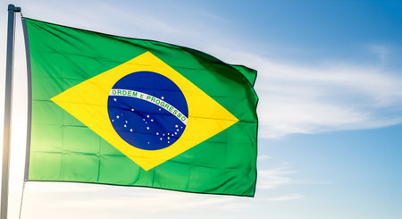 Brazilian flag is flying high showing patriotism pride colors bright clear blue sky. Representing Brazil national identity culture history strength. Seen from below fluttering representing unity progress independence symbolizing country spirit. Flag is bright sunlight blue sky behind.の素材