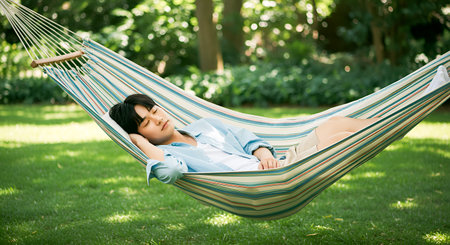 Relaxing hammock nap featuring a young man resting in hammock for a serene outdoor scene. Peaceful afternoon slumber with casual comfort in hammock set in garden. Sleep and daytime napping in natural surroundings. Emphasis on relaxation and tranquil setting. Hammock in a garden scene with tranquil rest.の素材