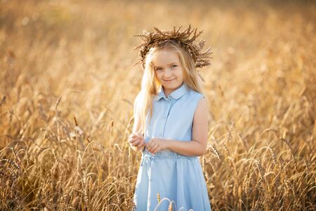 A little blonde girl is standing in a wheat field in summer with spikelets in her hands and a wreath on her head.の写真素材
