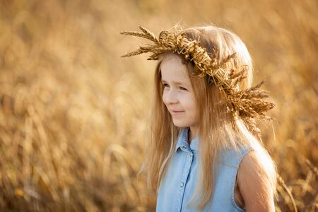 A little blonde girl is standing in a wheat field in summer and a wreath on her head.の写真素材
