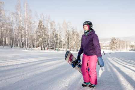 Girl in purple jacket and pink pants with snowboard in the hands of the uphill in winterの写真素材