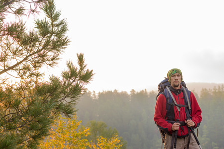 Man with backpack and trekking pole in bandana standing on a rock at dawn on a background autumn forest.の写真素材