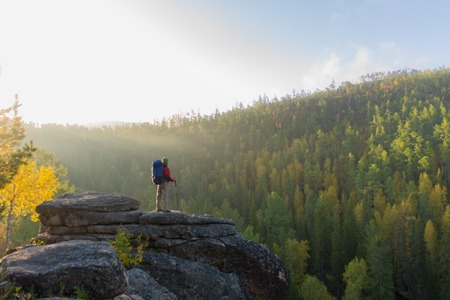 Man with backpack and trekking pole in bandana standing on a rock at dawn on a background autumn forest.の写真素材