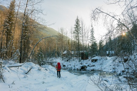 Woman with backpack walks along the mountain river in the snowdrifts in the winter forest at sunset.の写真素材